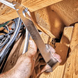 An electrician is using the Staple Shark to remove a cable staple from a wood beam. The staple shark has Rack-A-Tiers branding on it.